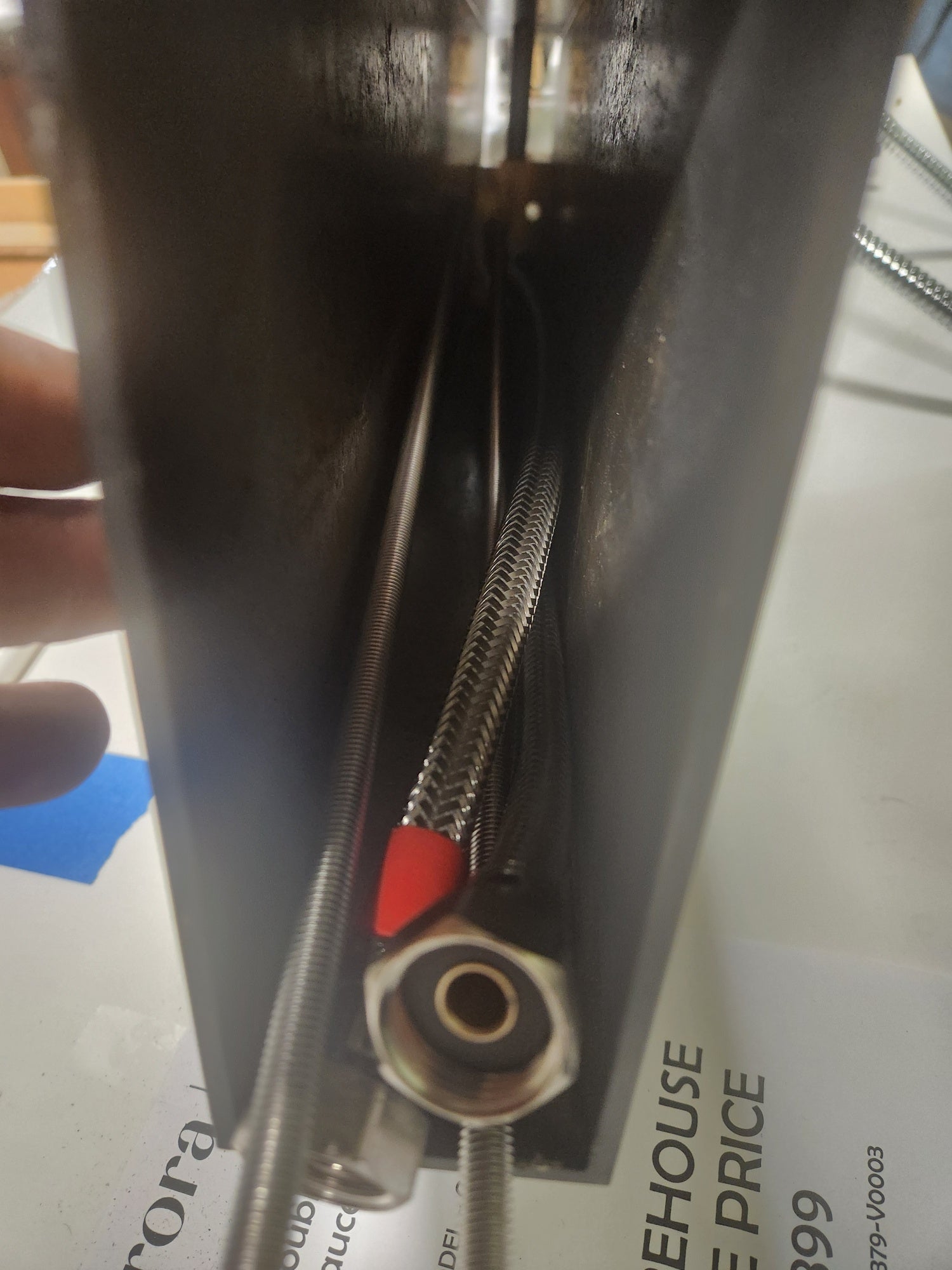 Close-up of a dark metal appliance or container showing a braided stainless steel water line with a red connector and a brass fitting, held by a hand over a printed paper background.