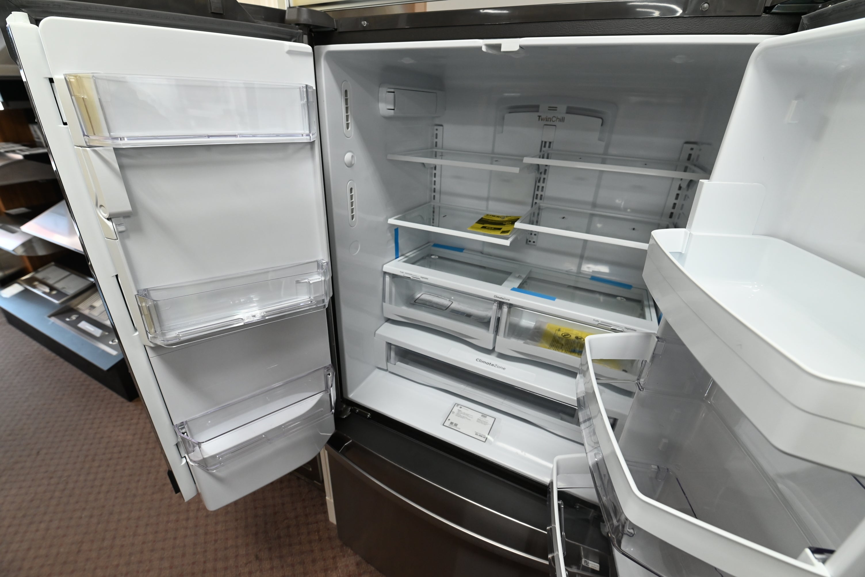 An open side-by-side refrigerator with its doors swung open, showing the white interior shelves, door bins, and lower freezer drawer. The appliance is photographed in a store or showroom setting with a brown carpeted floor visible.