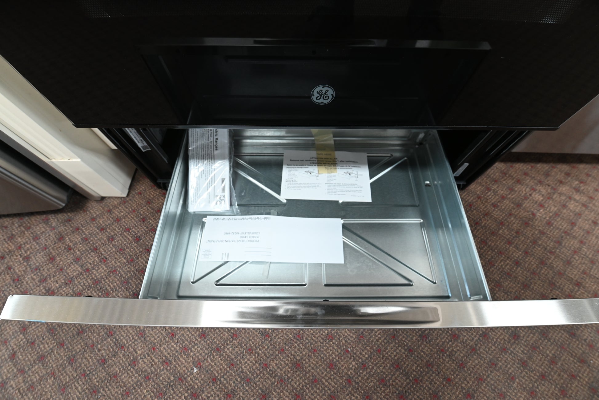 A GE built-in appliance drawer, likely a refrigerator or freezer storage compartment, shown open to reveal a stainless steel interior with paperwork inside. The appliance exterior is black, and the floor is carpeted.