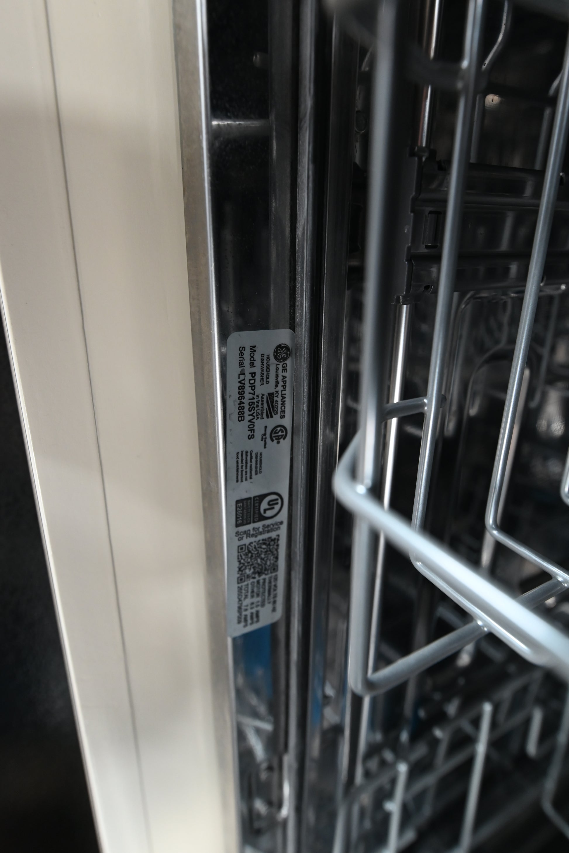 Close-up of the interior lower edge of a dishwasher, showing metal racks, a control panel area, and a product label with certification marks and a QR code.