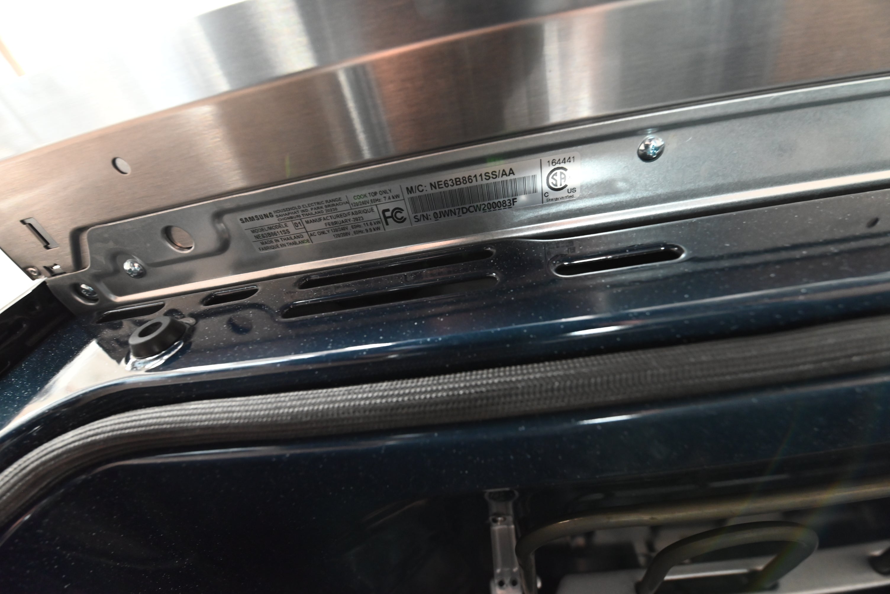 Close-up of the interior top edge of a dark appliance, showing a stainless steel panel, screws, a black rubber gasket, and a printed product label with regulatory text.