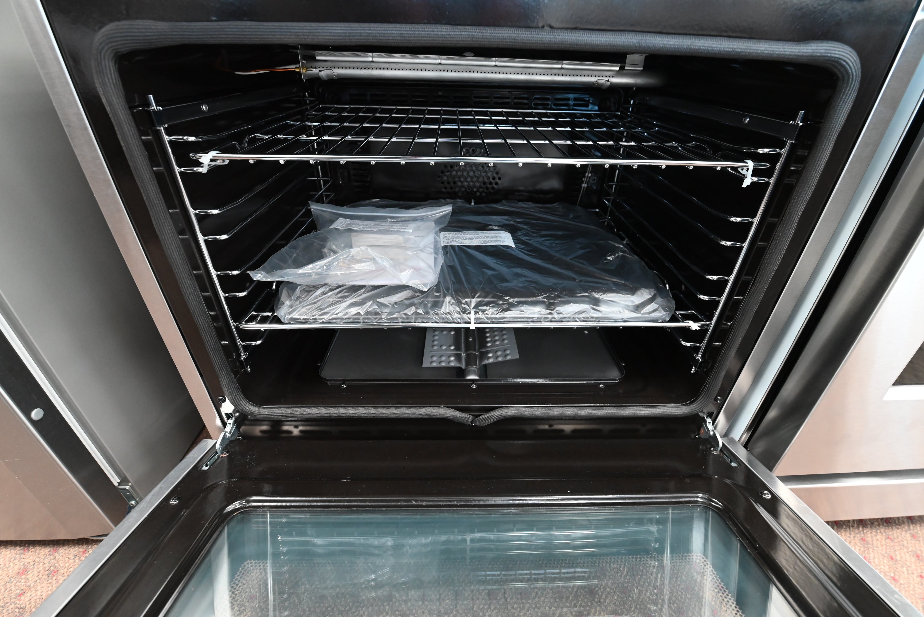 Open stainless steel built-in oven showing the interior racks, a plastic-wrapped item on the middle rack, and a black tray or insert on the lower rack.