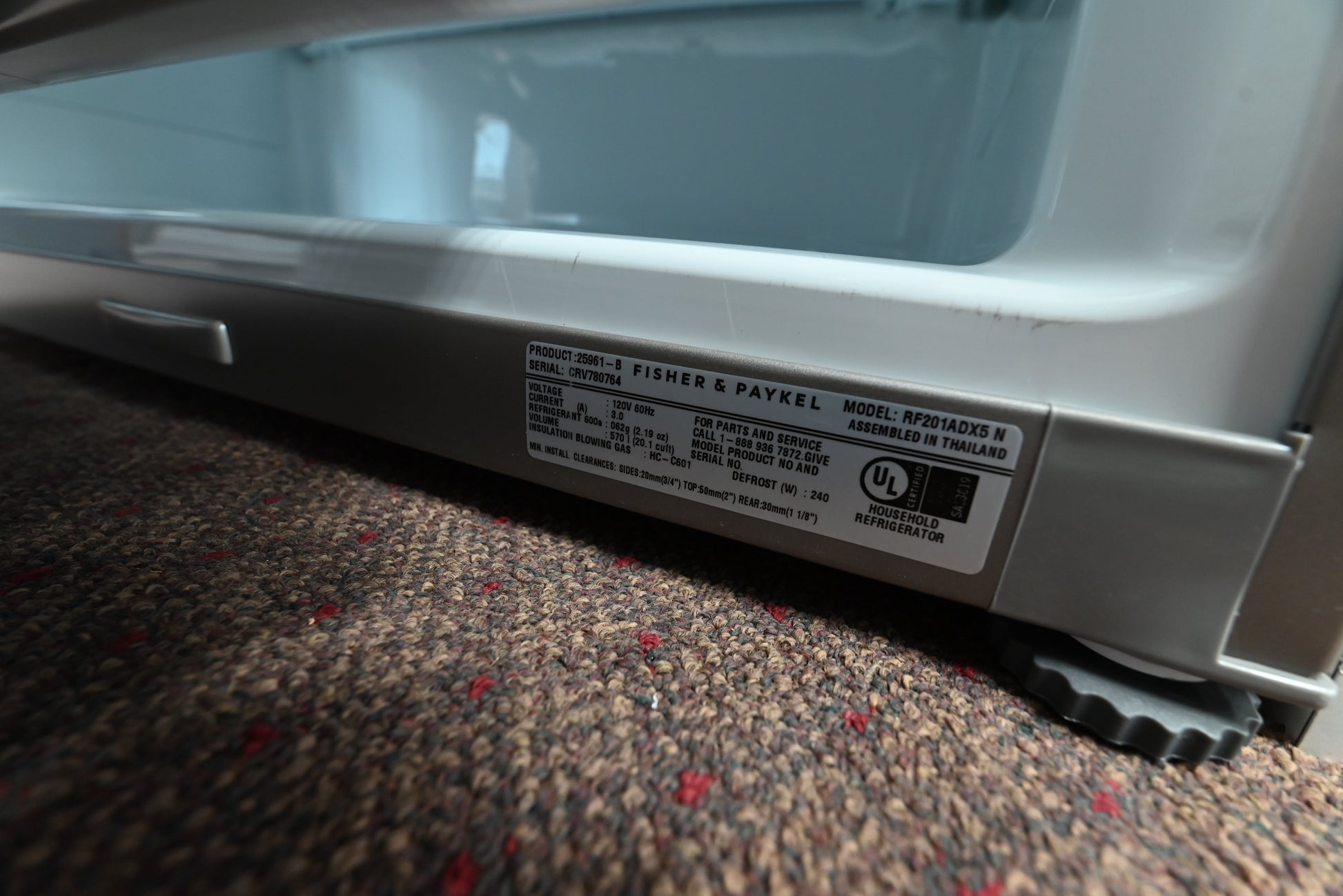 Close-up of the bottom edge of a silver appliance, showing a product label that reads Fisher & Paykel and a carpeted floor beneath it.