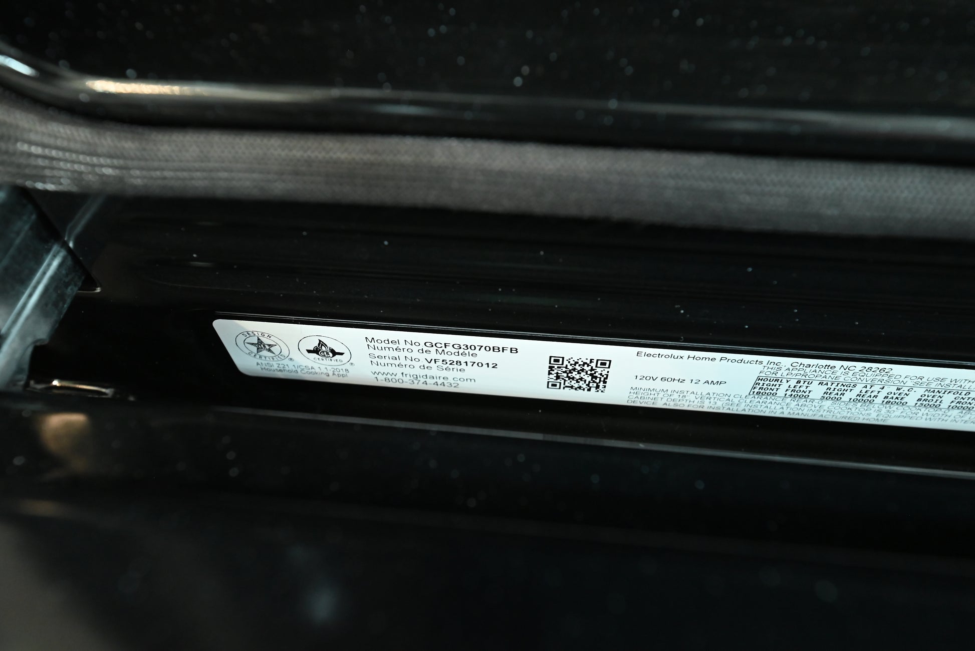 A close-up of a white product information label with a QR code and small printed text, attached to a black appliance or device.