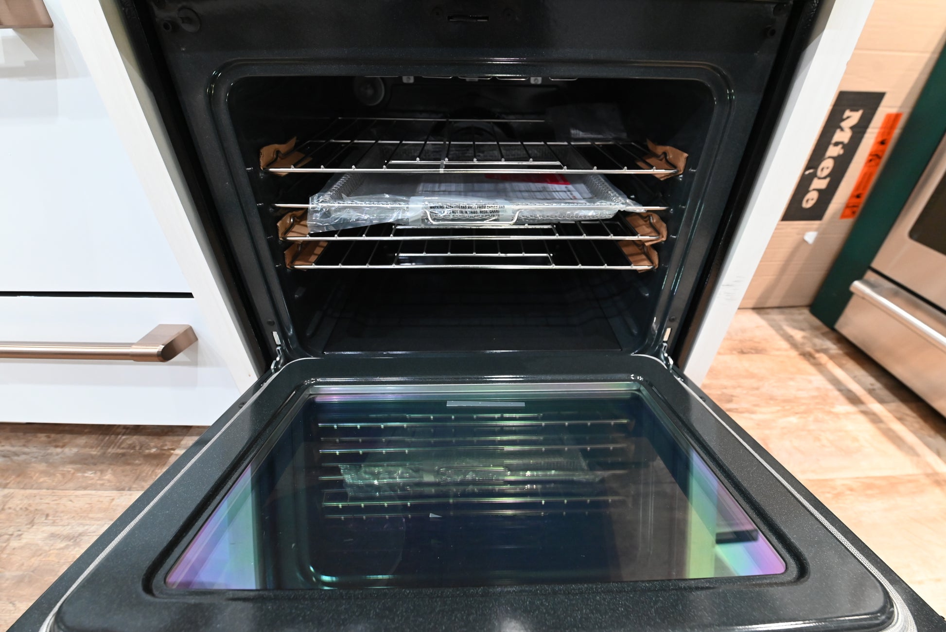 An open built-in oven with three racks inside, showing a plastic-wrapped item on one rack, set in a kitchen with white cabinetry and a wood floor.