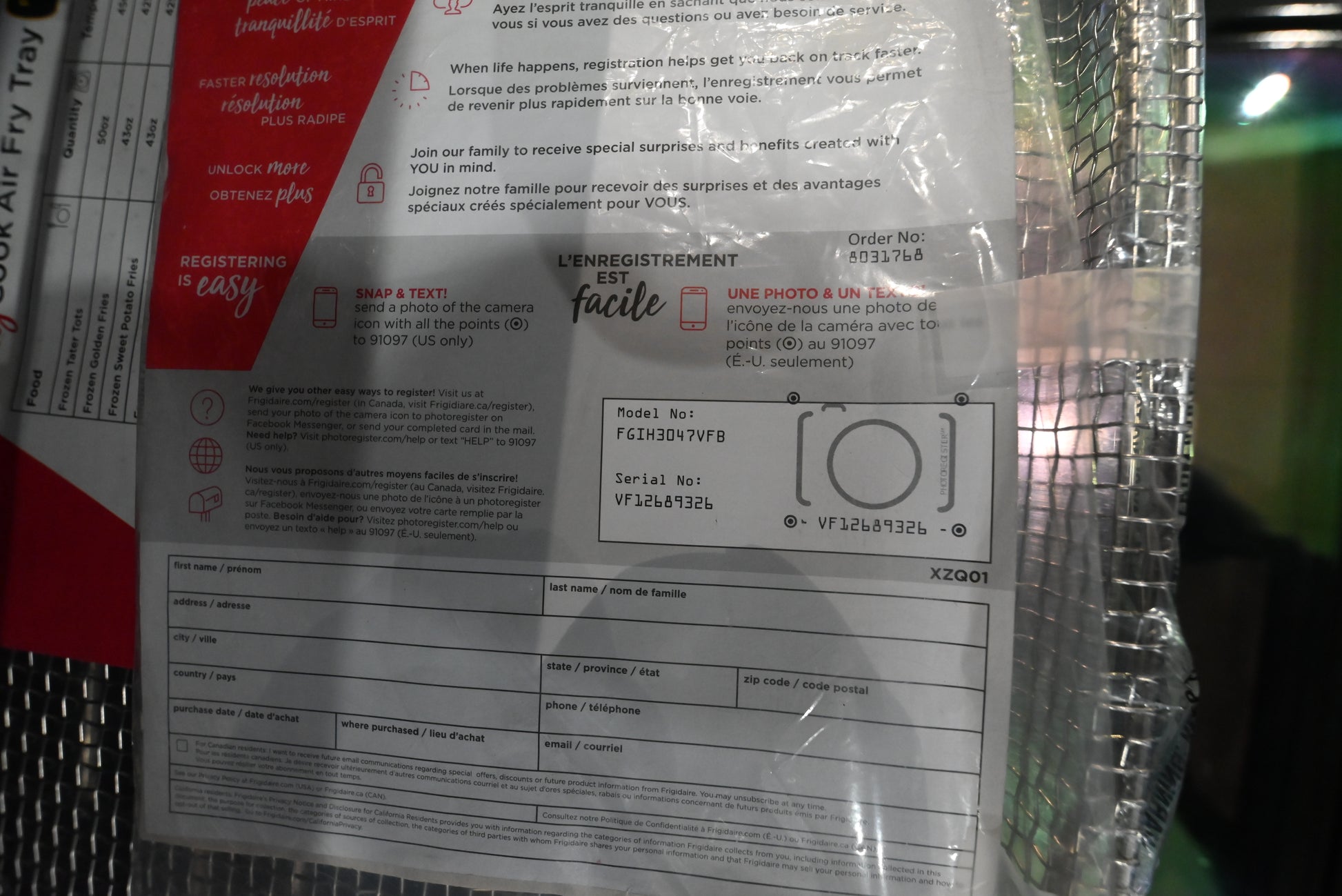 A close-up of a product's packaging label and manual, showing text in English and French, a barcode, and a camera icon diagram. The product is wrapped in clear plastic and sits on a metallic mesh shelf.
