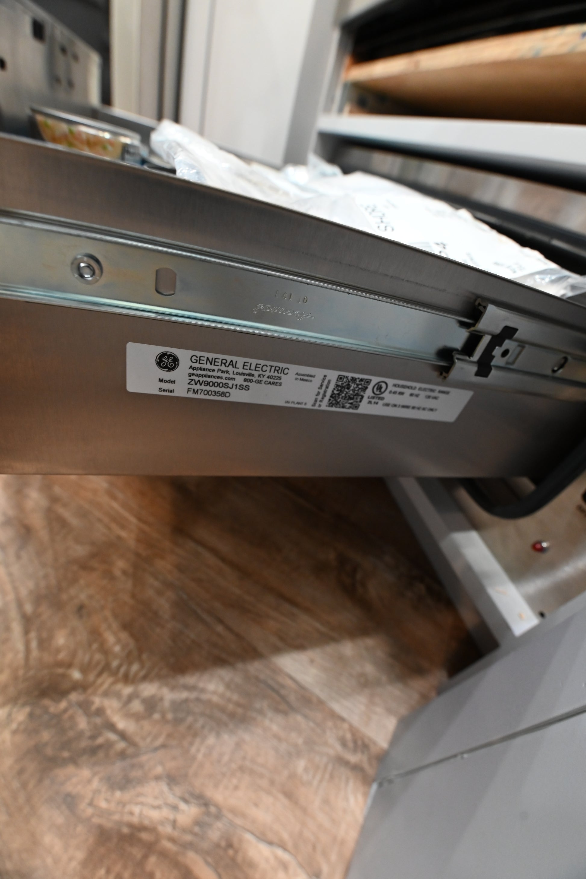 Close-up of a stainless steel appliance drawer or panel with a GE label sticker, shown in a real indoor setting with wood flooring visible.