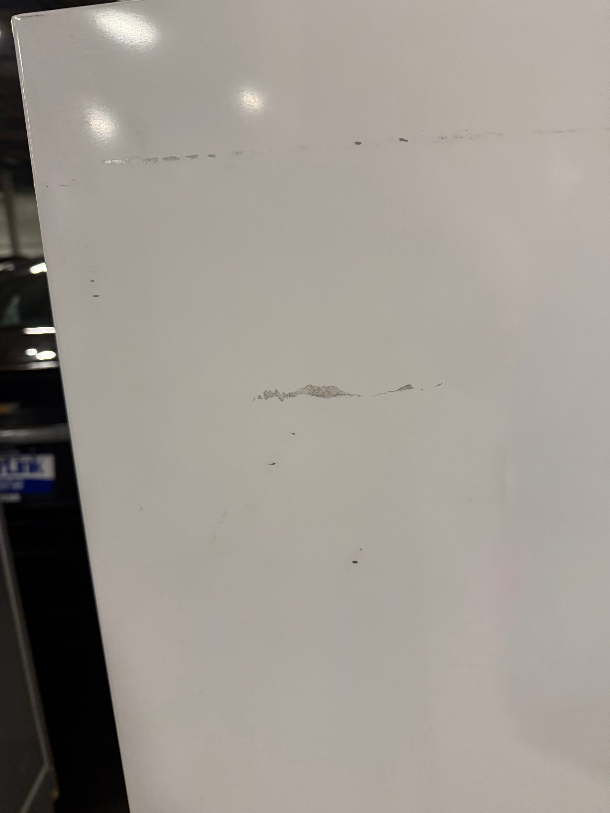 A close-up of a white panel or board with a few small scratches and marks, photographed indoors with some reflections and a dark background visible on the left.