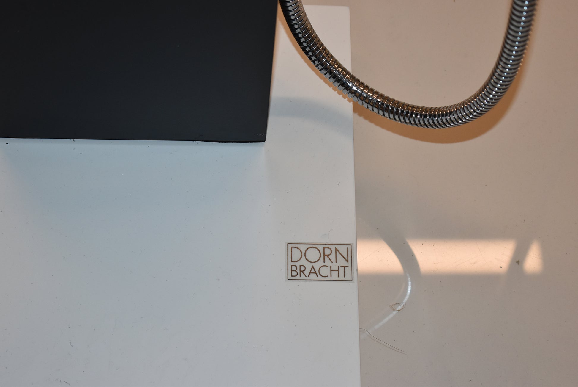 Close-up of a white wall or panel with a small square DORN BRACHT logo plaque, a black rectangular object at the top left, and a coiled metal hose running across the upper right.