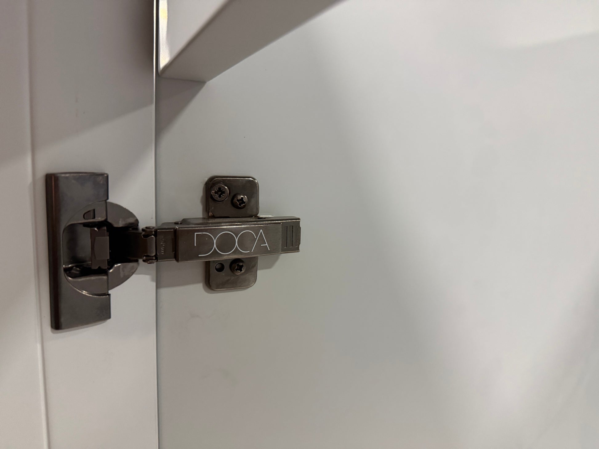 A close-up of a DOCA brand metal cabinet hinge mounted on a white cabinet door next to a shelf.