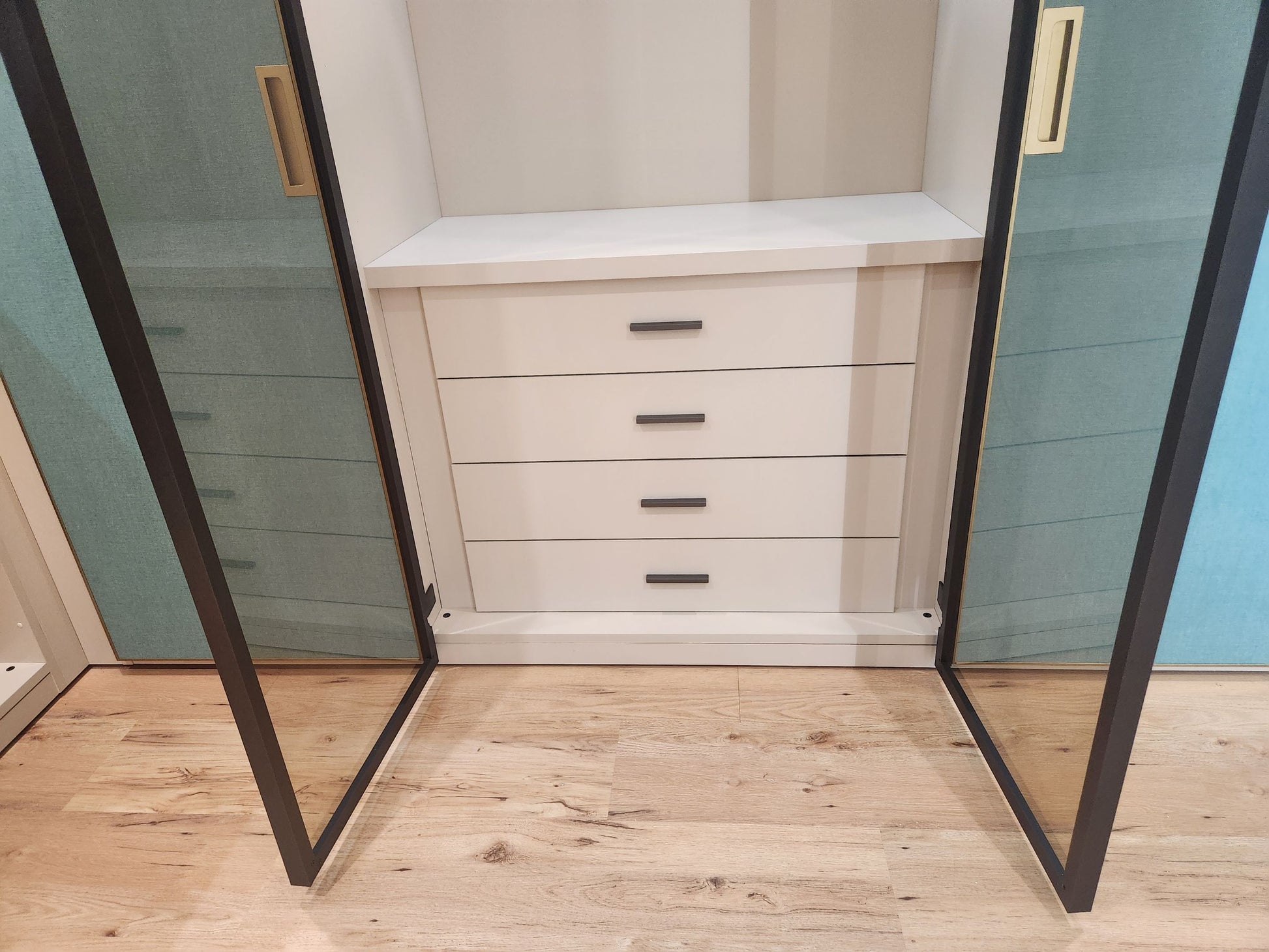 A white three-drawer storage cabinet with slim black handles, set inside a walk-in closet with open glass doors and light wood flooring.