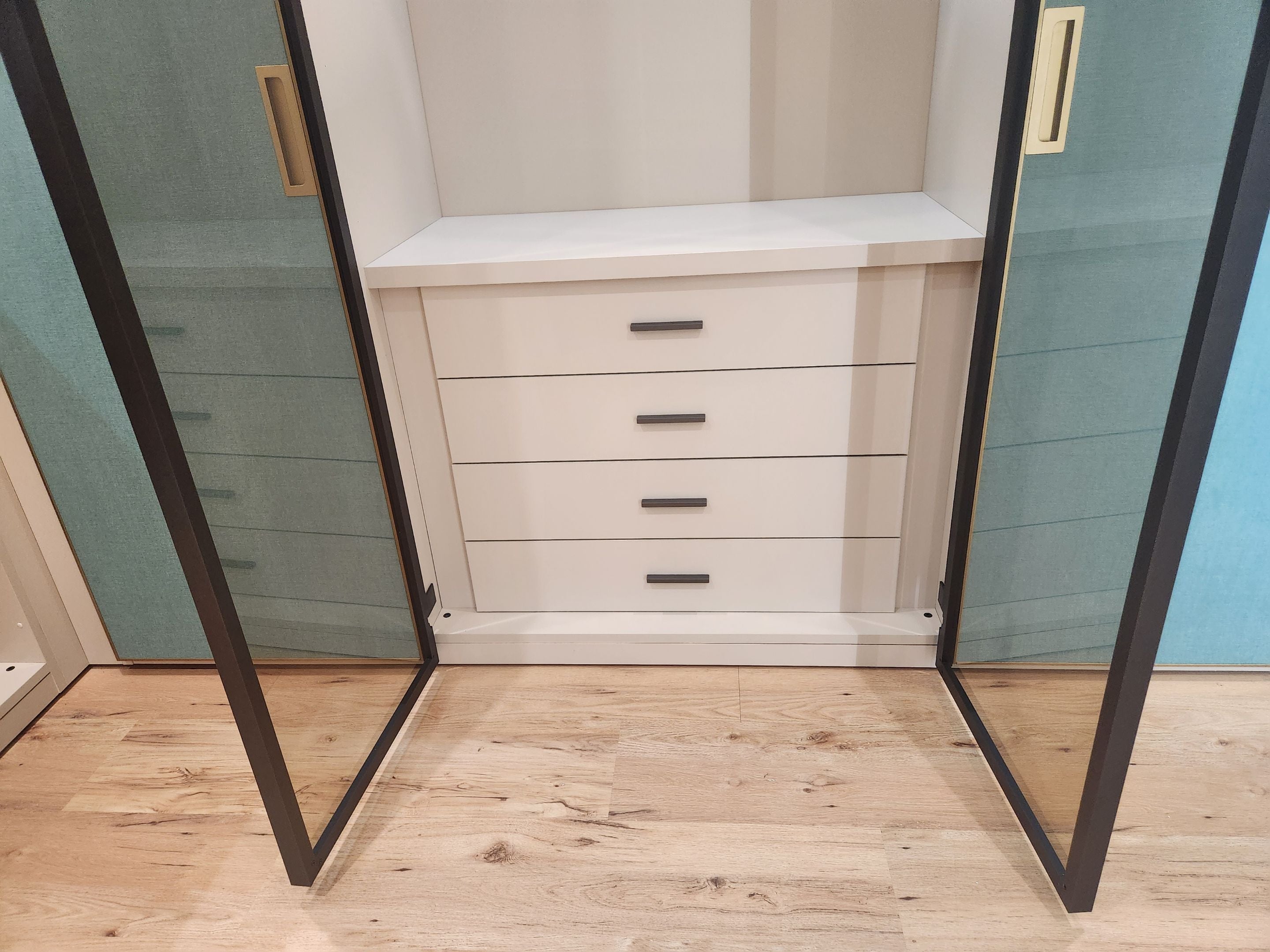 A white three-drawer storage cabinet with slim black handles, set inside a walk-in closet with open glass doors and light wood flooring.