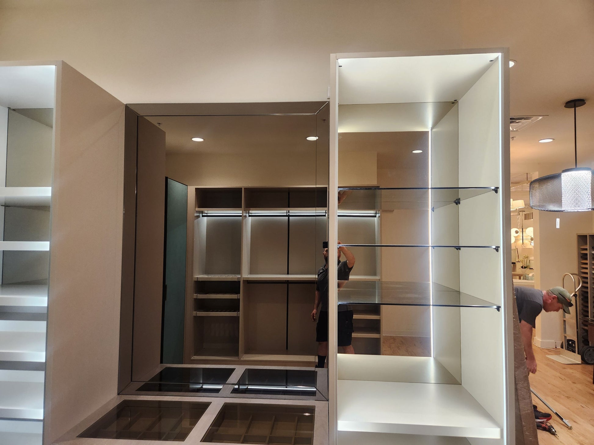 A large built-in walk-in closet system with illuminated white shelving and glass shelves, shown in a showroom or installation setting. A person is visible standing inside the closet, and another person is bent over on the right side.