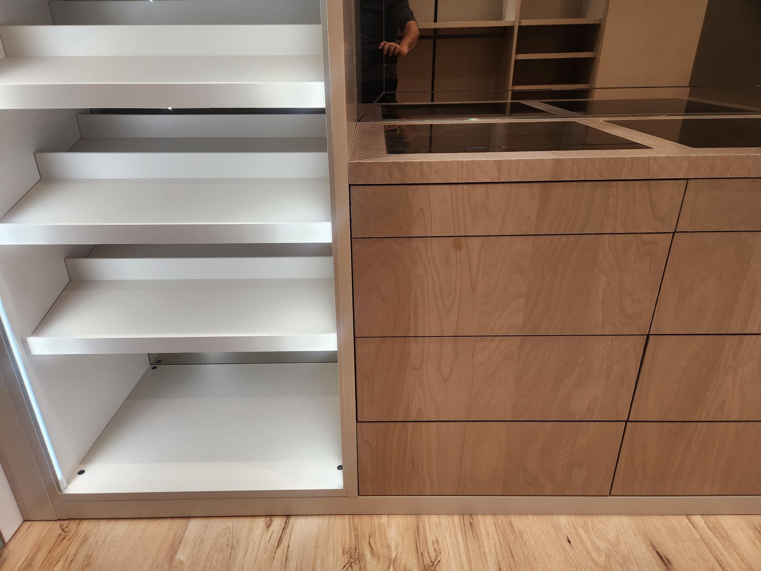 A modern built-in wardrobe or storage cabinet with white pull-out shelves on the left and light wood drawers on the right, installed in a room with a wood-look floor. A person is faintly visible in the reflection above the counter.