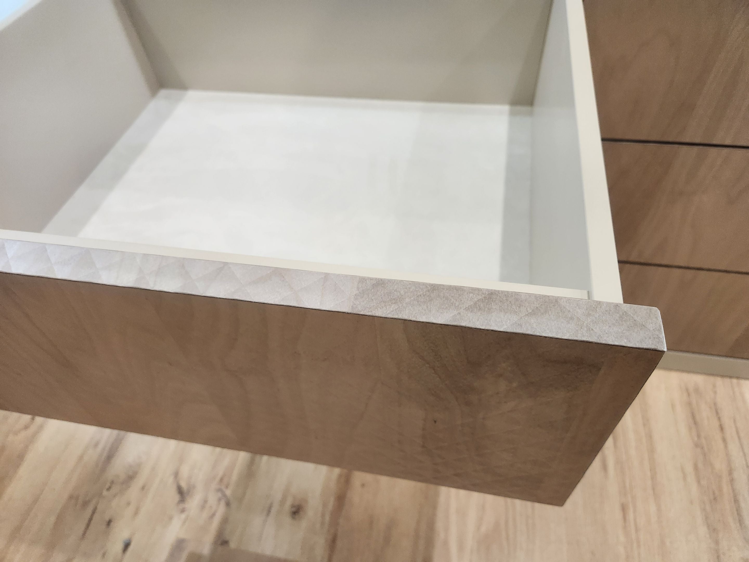 An open wooden drawer with a light wood grain exterior and a white interior, shown on a light wood floor next to matching drawers.