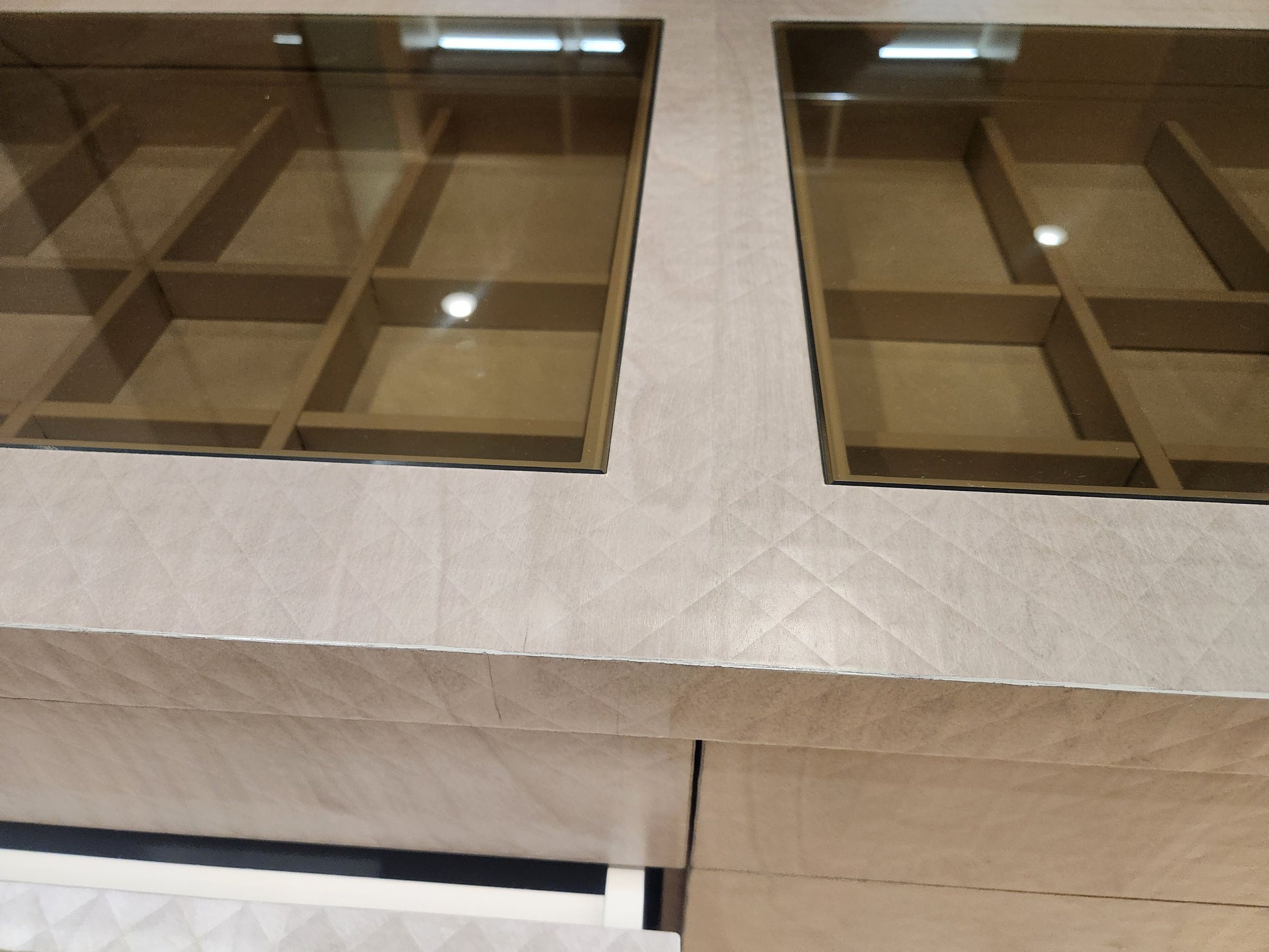 Close-up of a modern vanity or cabinet interior with two rectangular glass-front storage compartments set into a light beige textured stone or laminate front, with warm wood-toned shelves visible behind the glass.