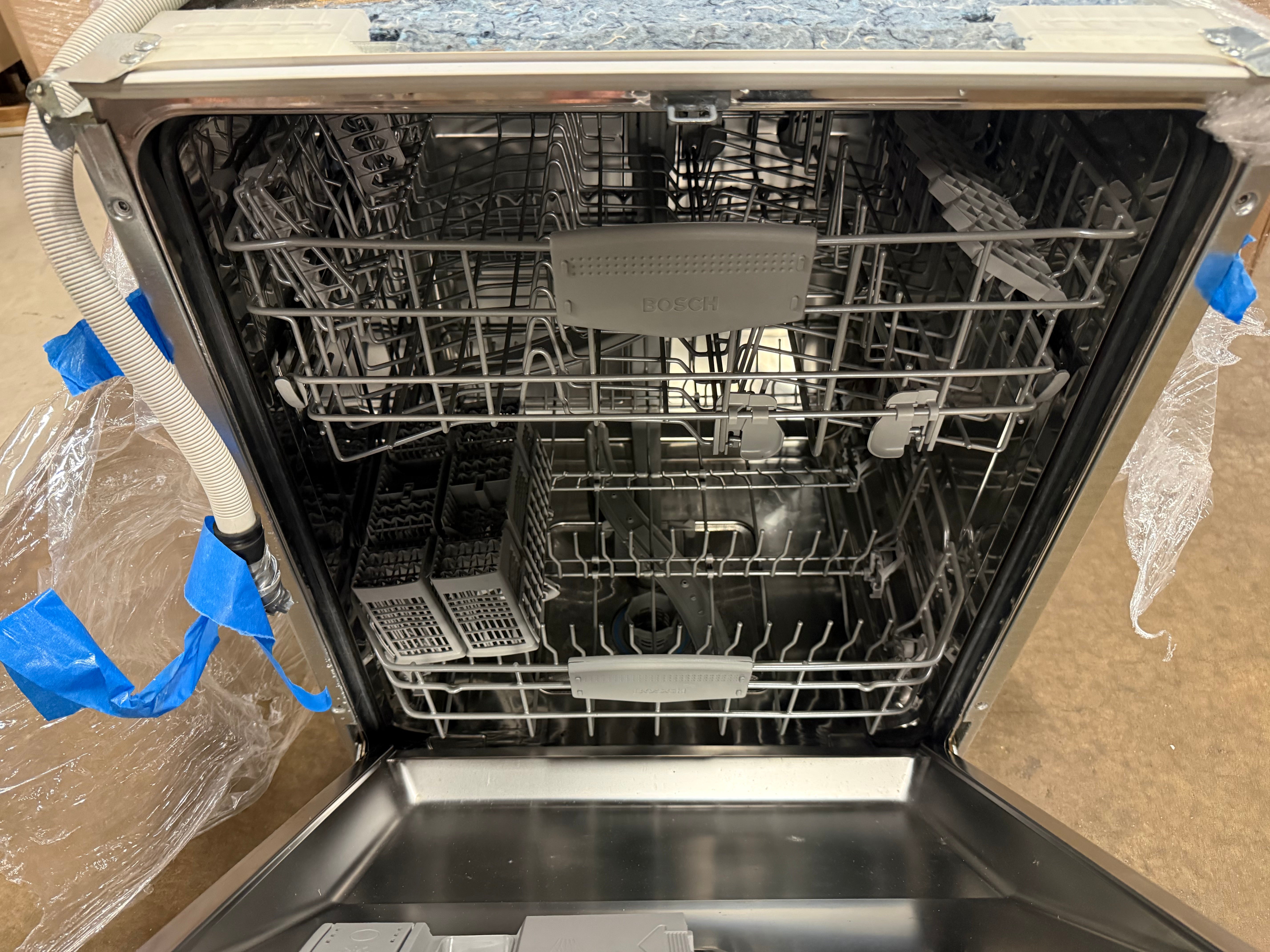 Open stainless steel dishwasher showing the interior racks, detergent drawer area, and protective blue tape and plastic wrapping around the appliance.
