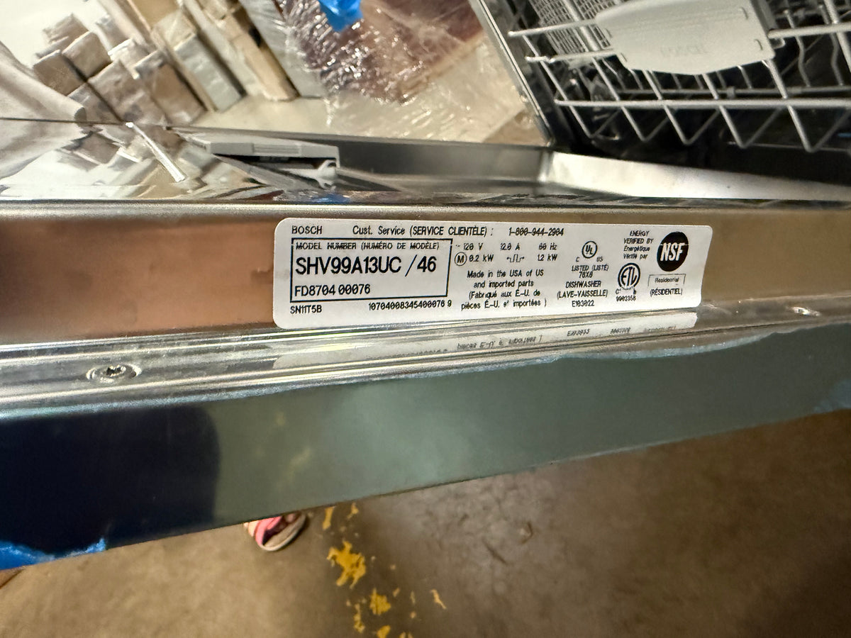 A close-up of a Bosch dishwasher's metal control panel edge with a white service information label showing model SHV99A1UC / 46 and Bosch branding, photographed in a warehouse or appliance storage setting.
