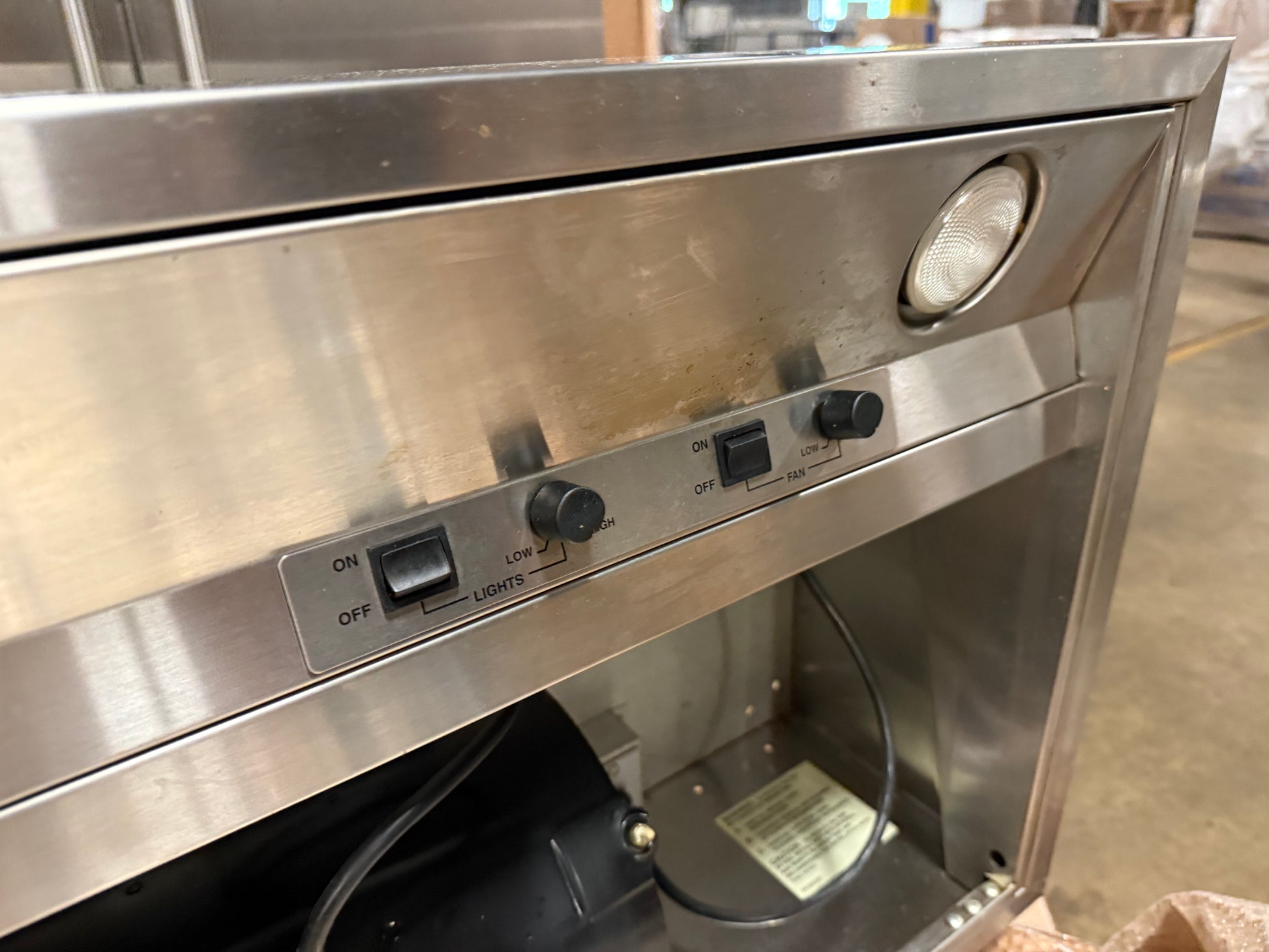 A stainless steel commercial range hood or vent cover with front-mounted control switches, a round light, and an open lower compartment showing internal wiring.
