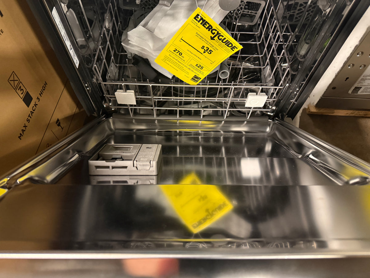 Open stainless steel dishwasher showing the interior racks, yellow EnergyGuide labels, and a cardboard box on the left side.