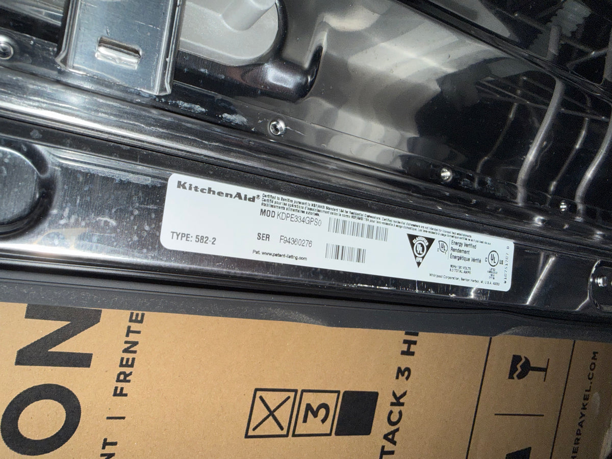 A close-up of a KitchenAid appliance label sticker on a metallic chrome surface, with part of a brown cardboard shipping box visible below.