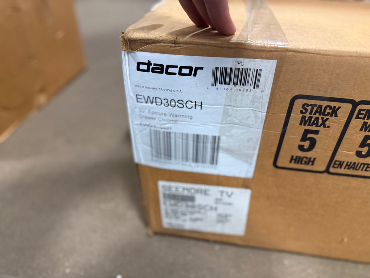 A brown cardboard box with a white Dacor label reading EWD30SCH, held by a hand on a concrete floor. The box also has a black 'STACK MAX.