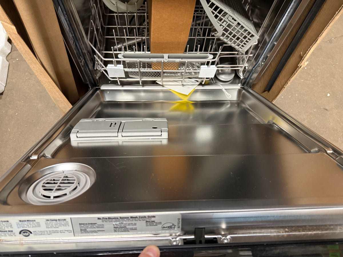 Open stainless steel dishwasher with the door swung open, showing the interior racks, detergent drawer, and door panel. A hand is visible at the bottom edge, and the appliance is photographed in a warehouse or storage setting with cardboard and concrete around it.