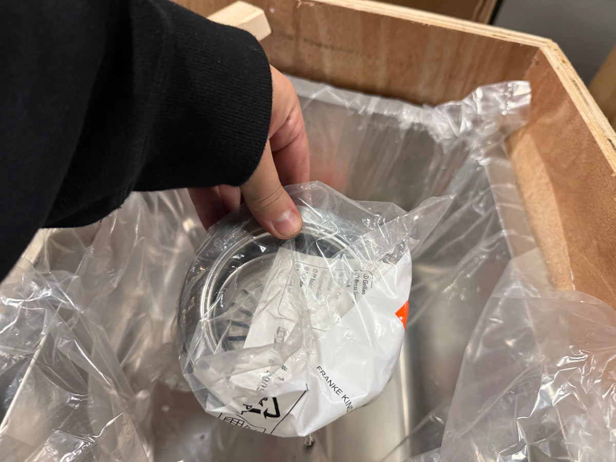 A hand wearing a black sleeve holds a packaged metal appliance part inside a cardboard shipping box with clear plastic wrapping.