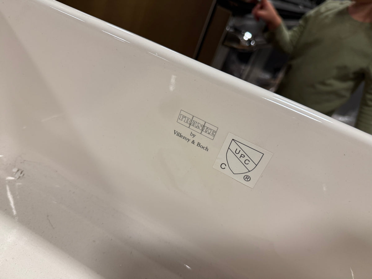 Close-up of the inside corner of a glossy white porcelain bathtub, showing a printed 'michelson by Vitrancy & Bosch' sticker and a 'CUPC' logo. The background shows a blurry kitchen environment with a person in an olive green shirt.