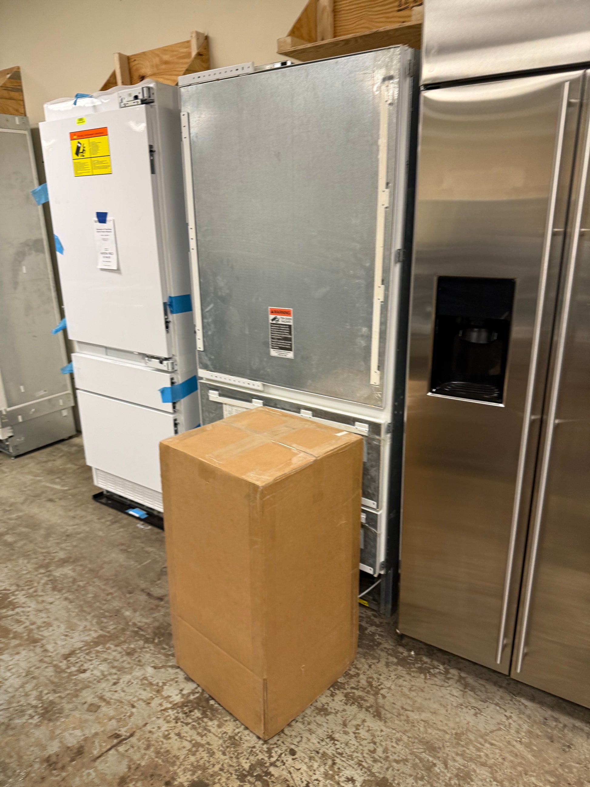 A stainless steel side-by-side refrigerator stands next to a white top-freezer refrigerator, with a large cardboard box placed in front of them on a concrete floor.