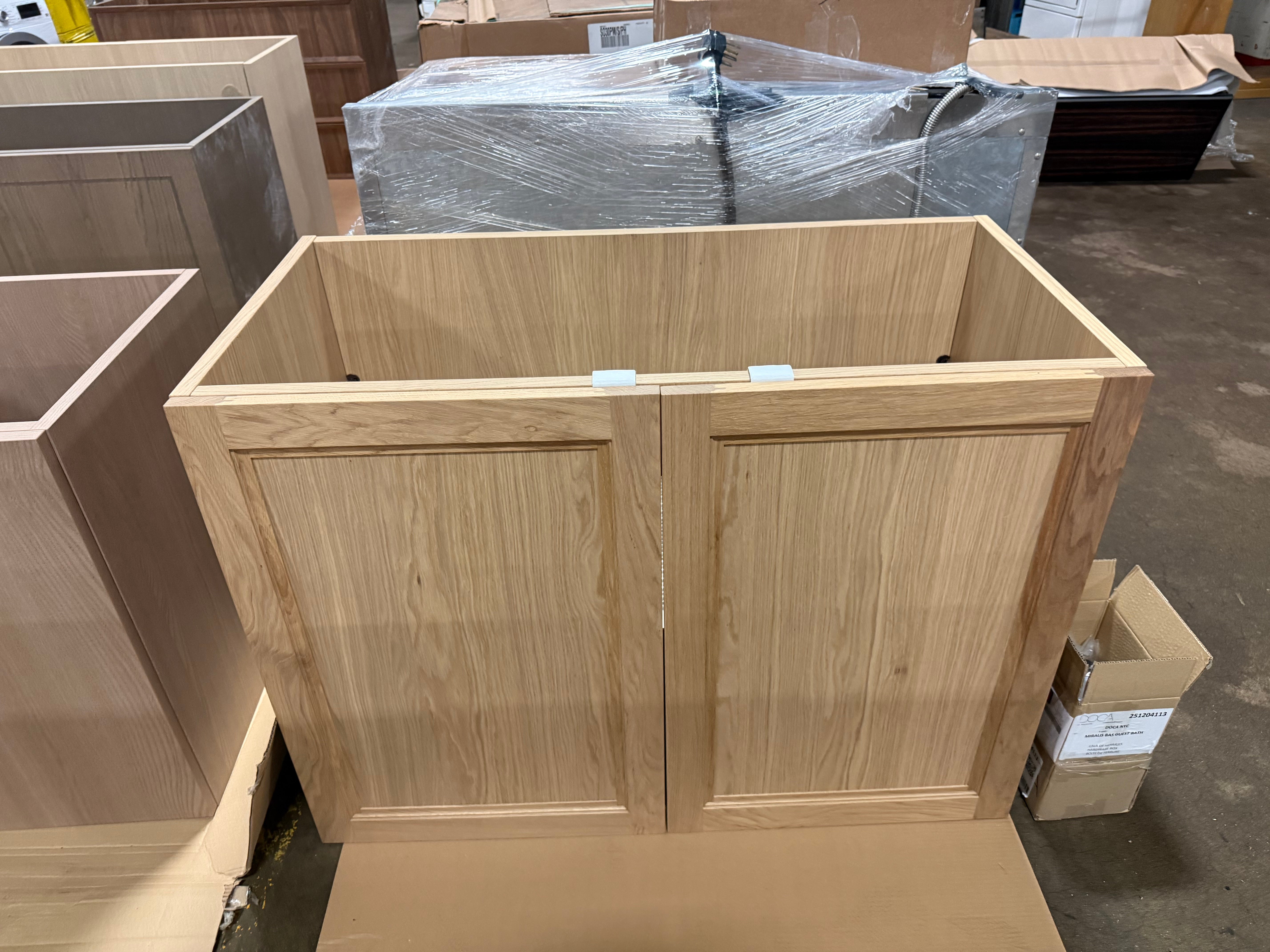 A light oak wood double cabinet base with paneled doors, shown in a warehouse setting with other furniture pieces and cardboard boxes around it.