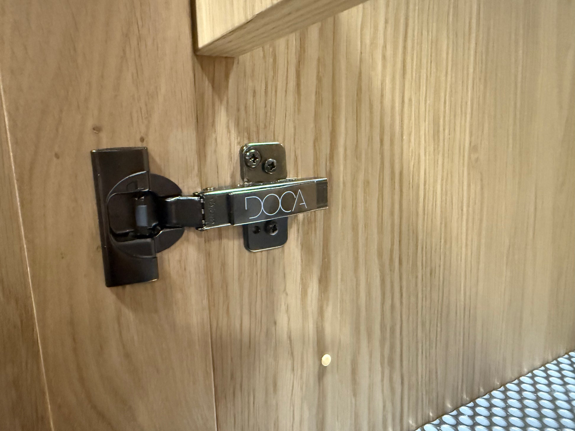 A close-up of a dark metal cabinet hinge mounted on a light wood grain door, with the brand name 'DOCA' visible on the hinge arm.