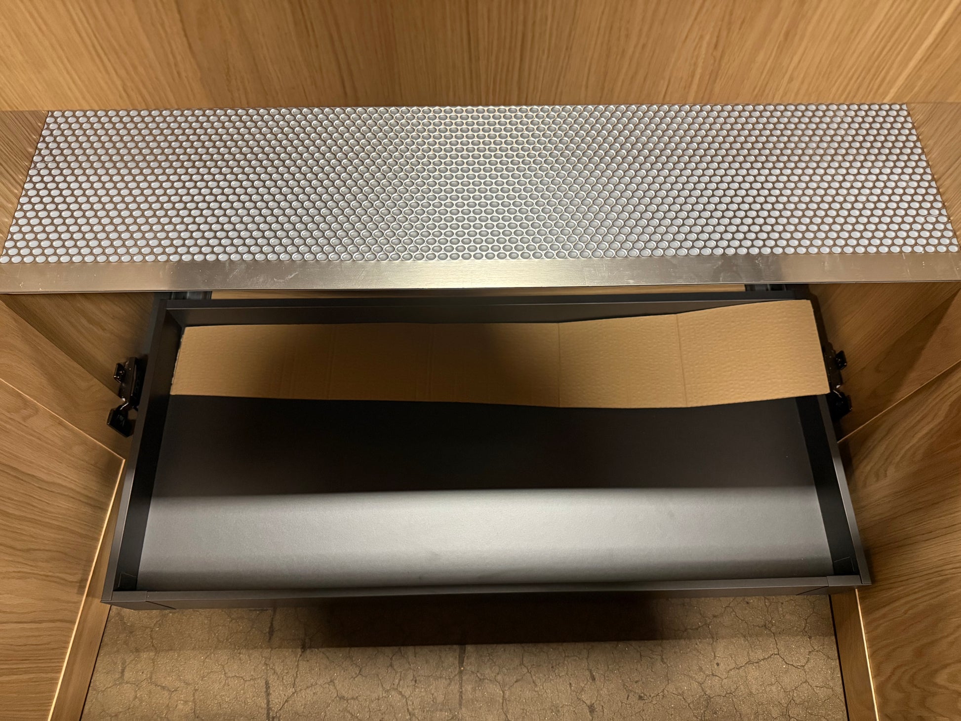 A close-up view of a metallic drawer or compartment with a textured silver top surface and a dark interior, set within a wood-toned cabinet or enclosure.