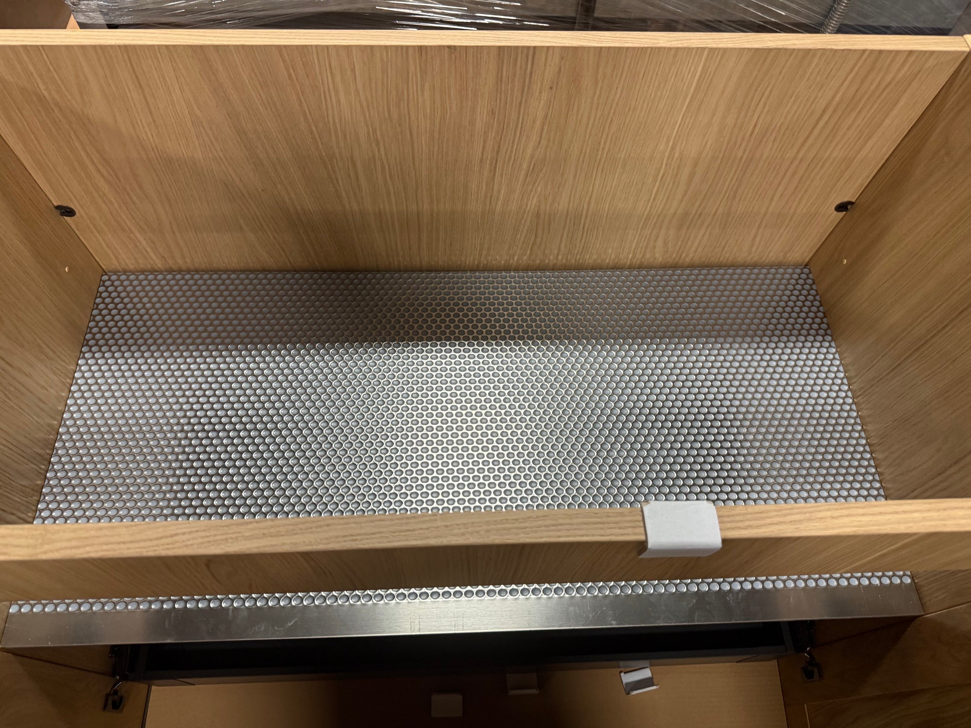 A close-up view inside a wooden cabinet or drawer showing a silver metal wire mesh shelf liner and a metal mounting track.