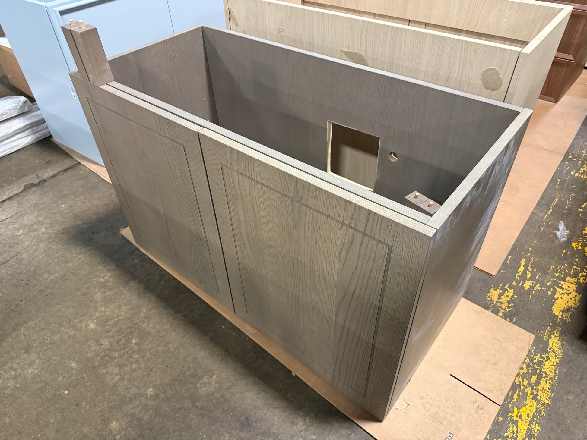 A large unfinished wooden cabinet or utility box with a woodgrain finish, photographed in a workshop or warehouse setting on a concrete floor.