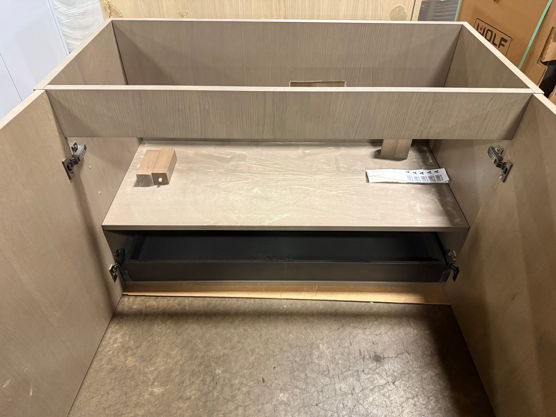 Open light wood cabinet or vanity base with a shelf and lower drawer compartment, photographed in a warehouse or showroom setting.