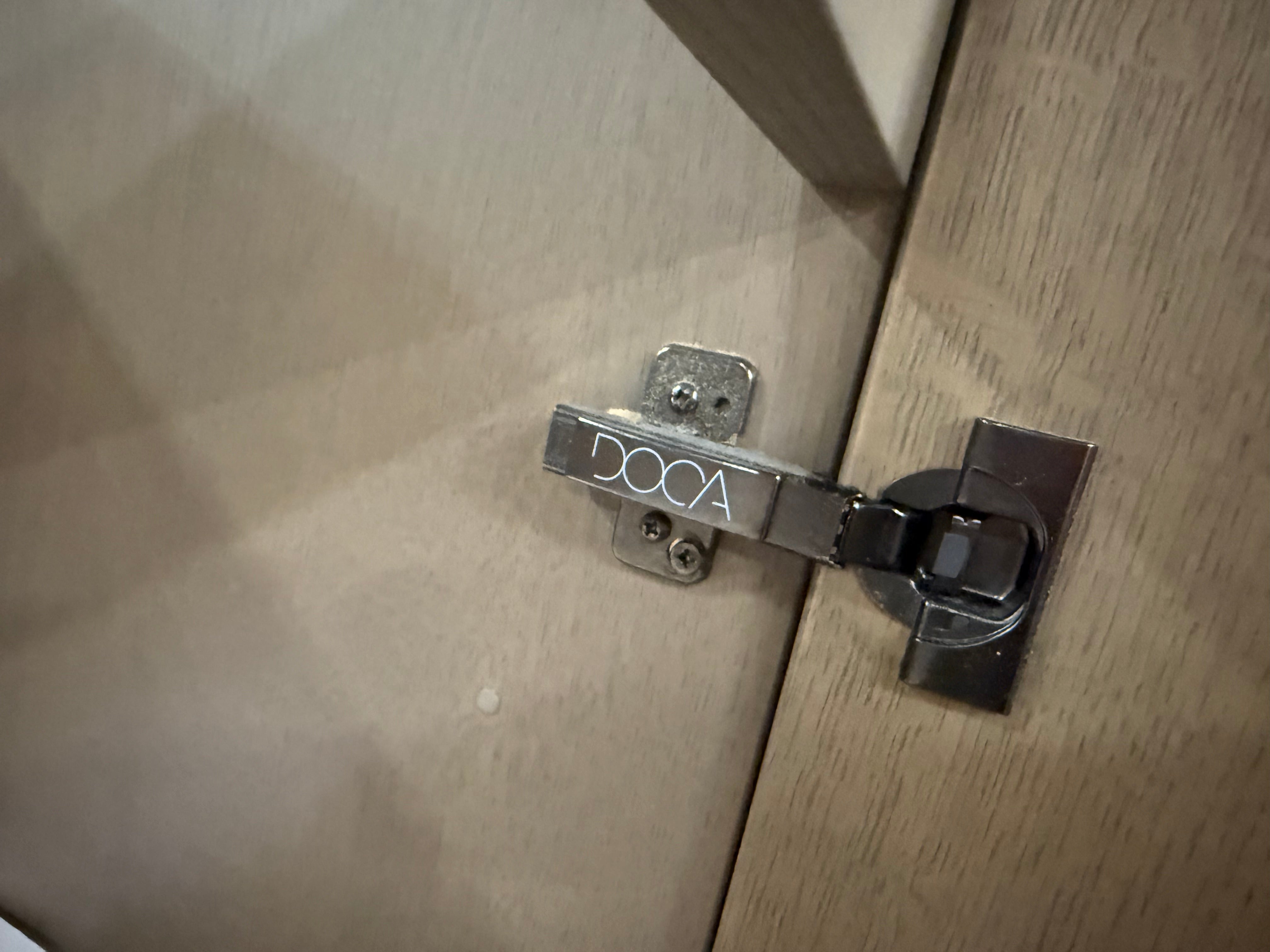 A close-up of a DOCA brand metal cabinet latch installed on a light wood door, showing the brand name engraved on the lever.