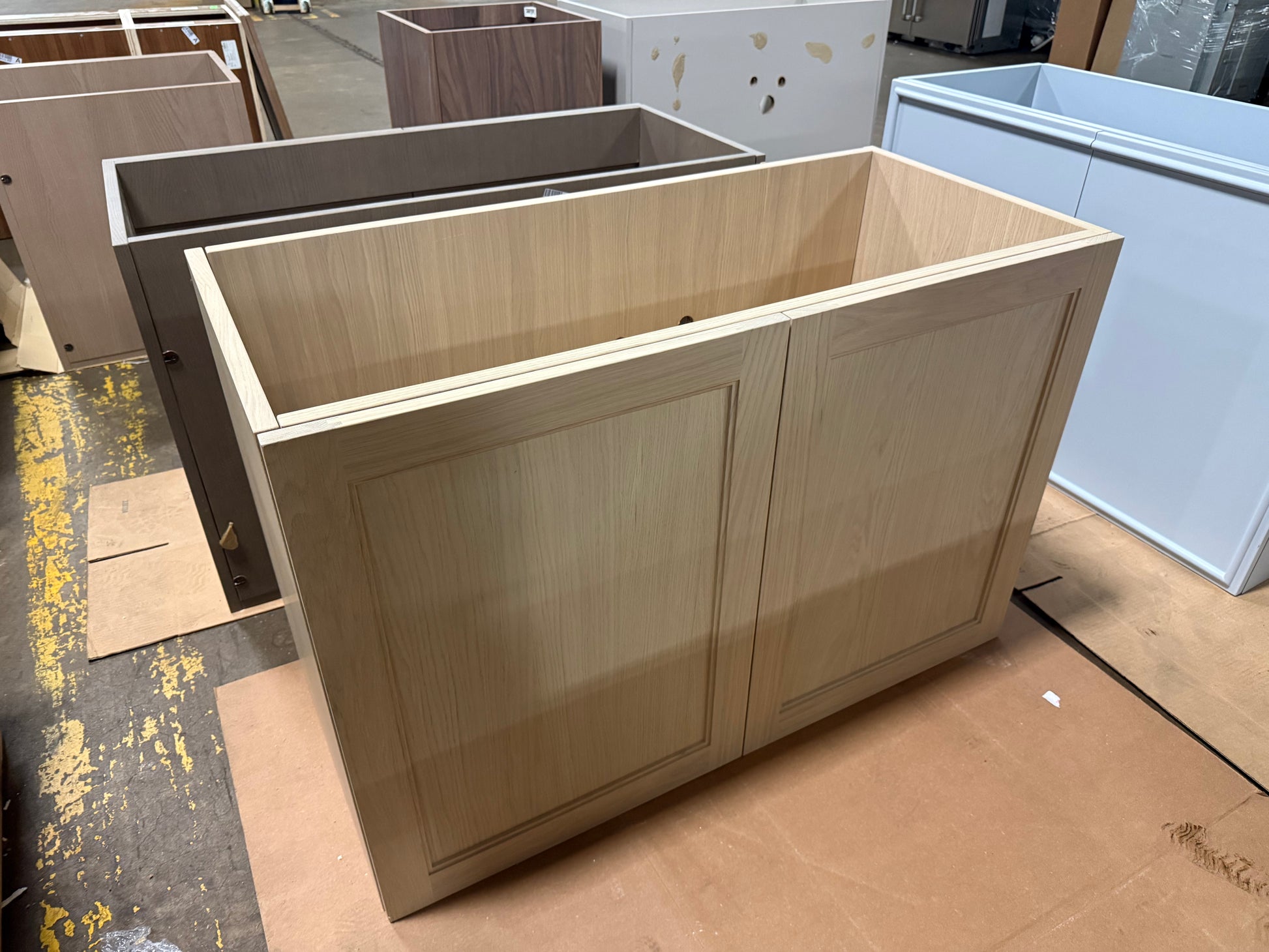A light natural wood double cabinet base, likely for a vanity or kitchen island, shown in a workshop setting with other unfinished cabinetry pieces around it.