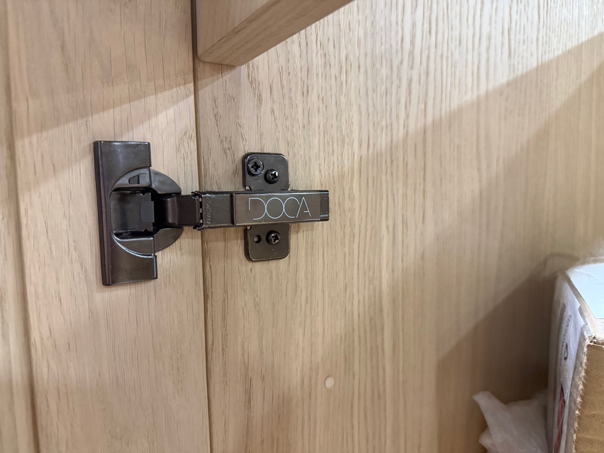 A close-up of a dark metal hinge mounted on a light wood cabinet door, with the brand name 'DOC A' visible on the hinge arm.