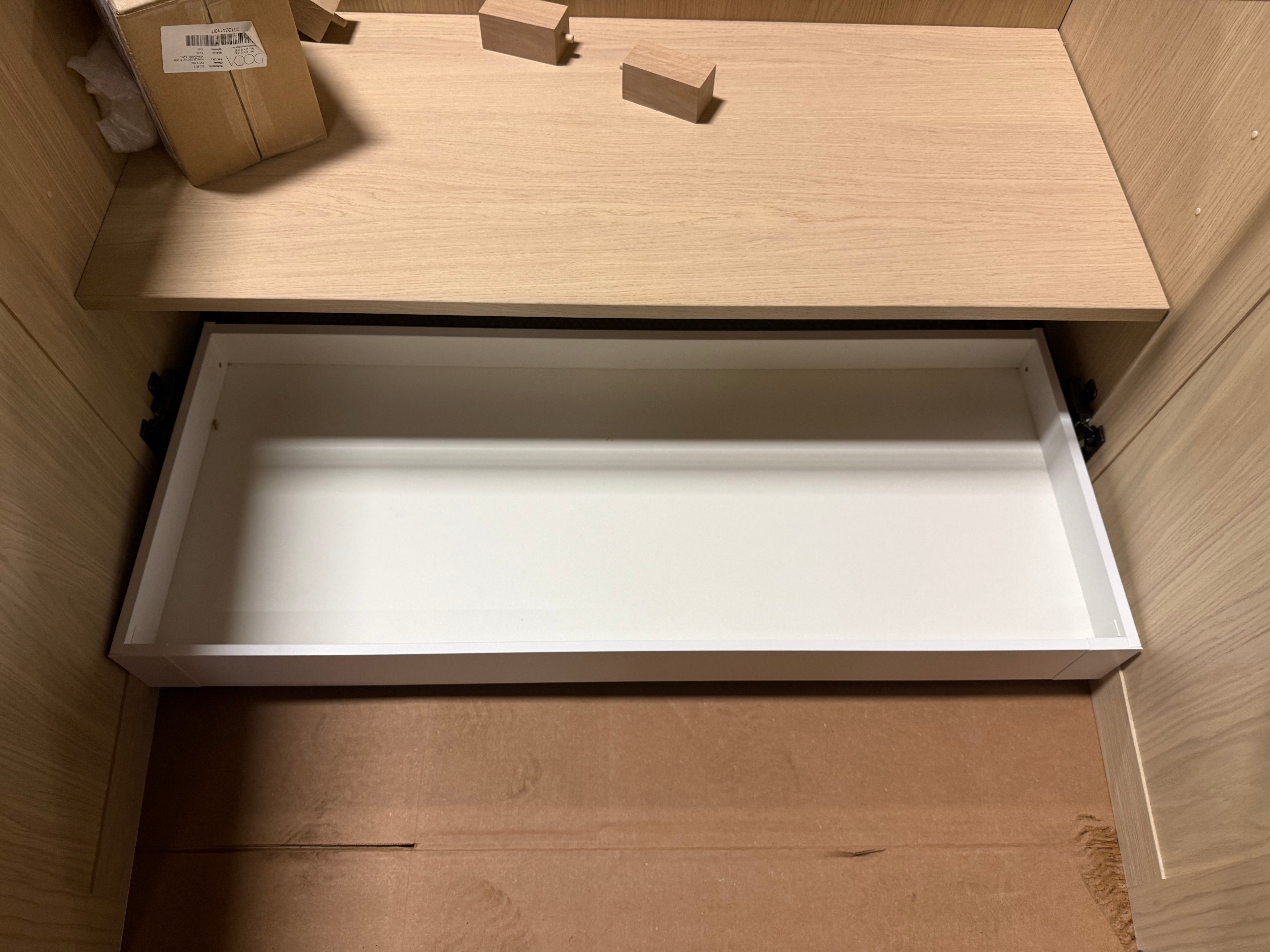 A white drawer pull-out tray inside a light wood cabinet or shelf, with cardboard packaging pieces visible nearby.