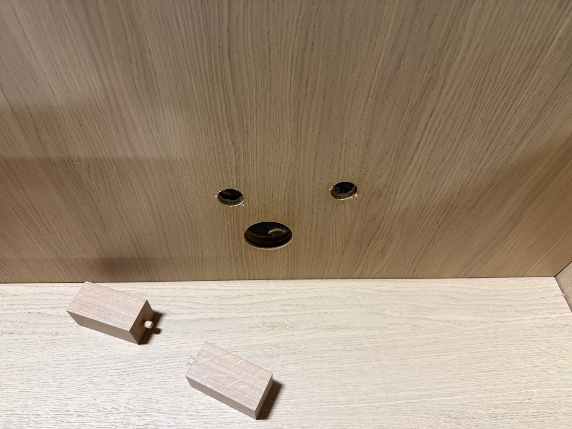 Close-up of a light wood cabinet interior showing three pre-drilled holes on the back panel and two small wooden dowel pieces on the shelf surface.