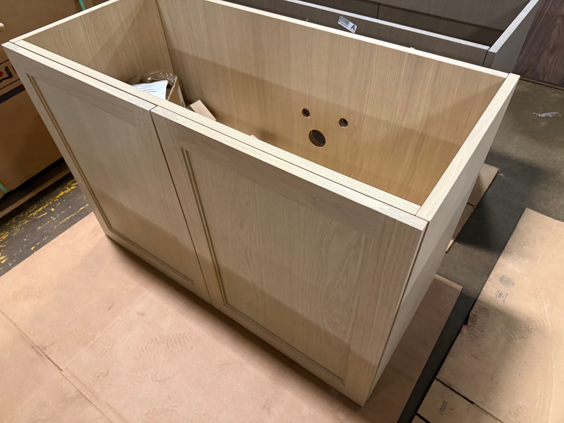 A light natural wood cabinet base with two paneled doors and a raised front apron, photographed in a workshop or warehouse setting on cardboard flooring.