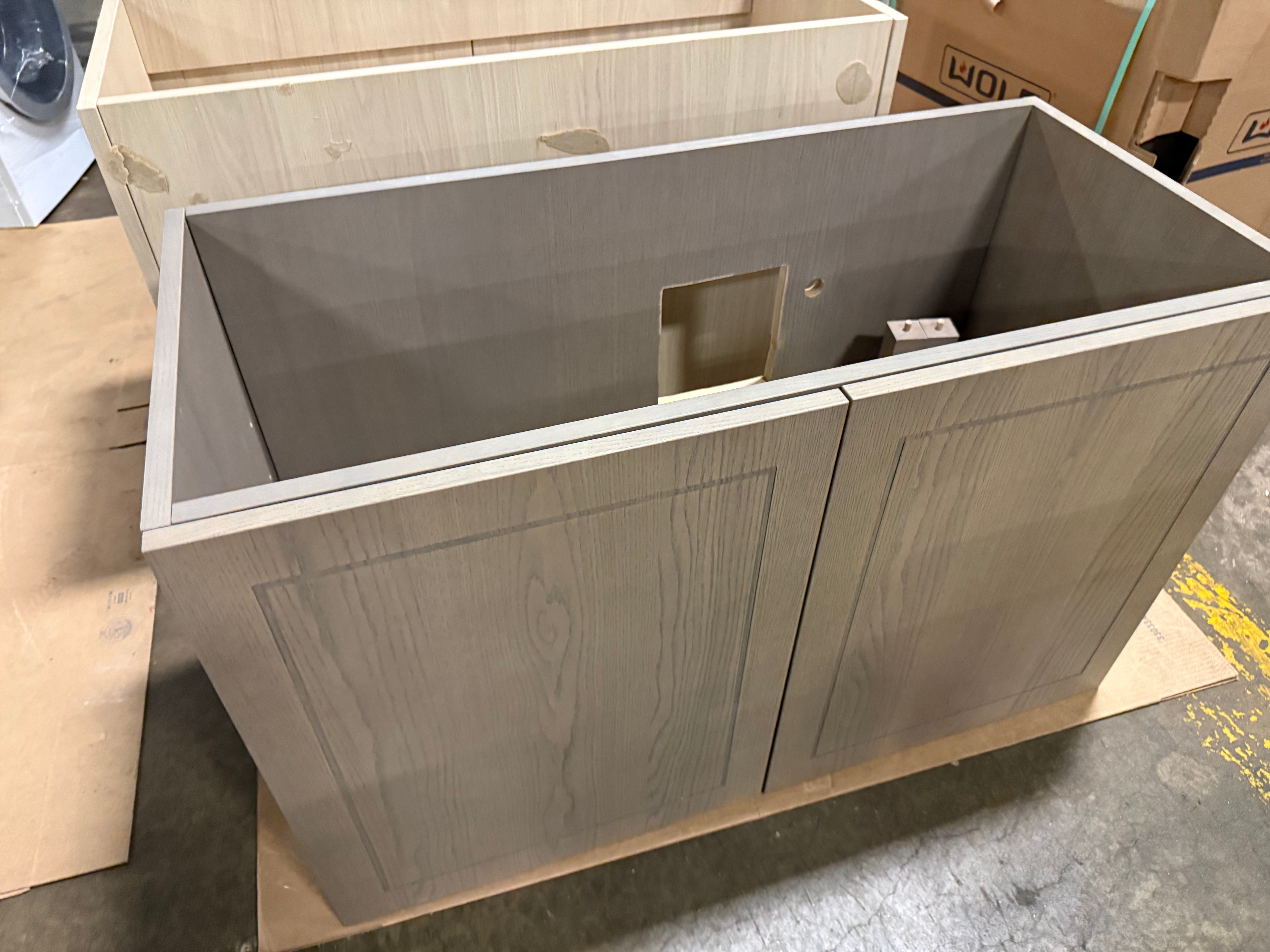 A light gray wood-grain cabinet base with two front doors and a recessed center panel, shown empty with its interior opening visible. The cabinet sits on a wooden support board in a warehouse or workshop setting with cardboard boxes and concrete floor visible.