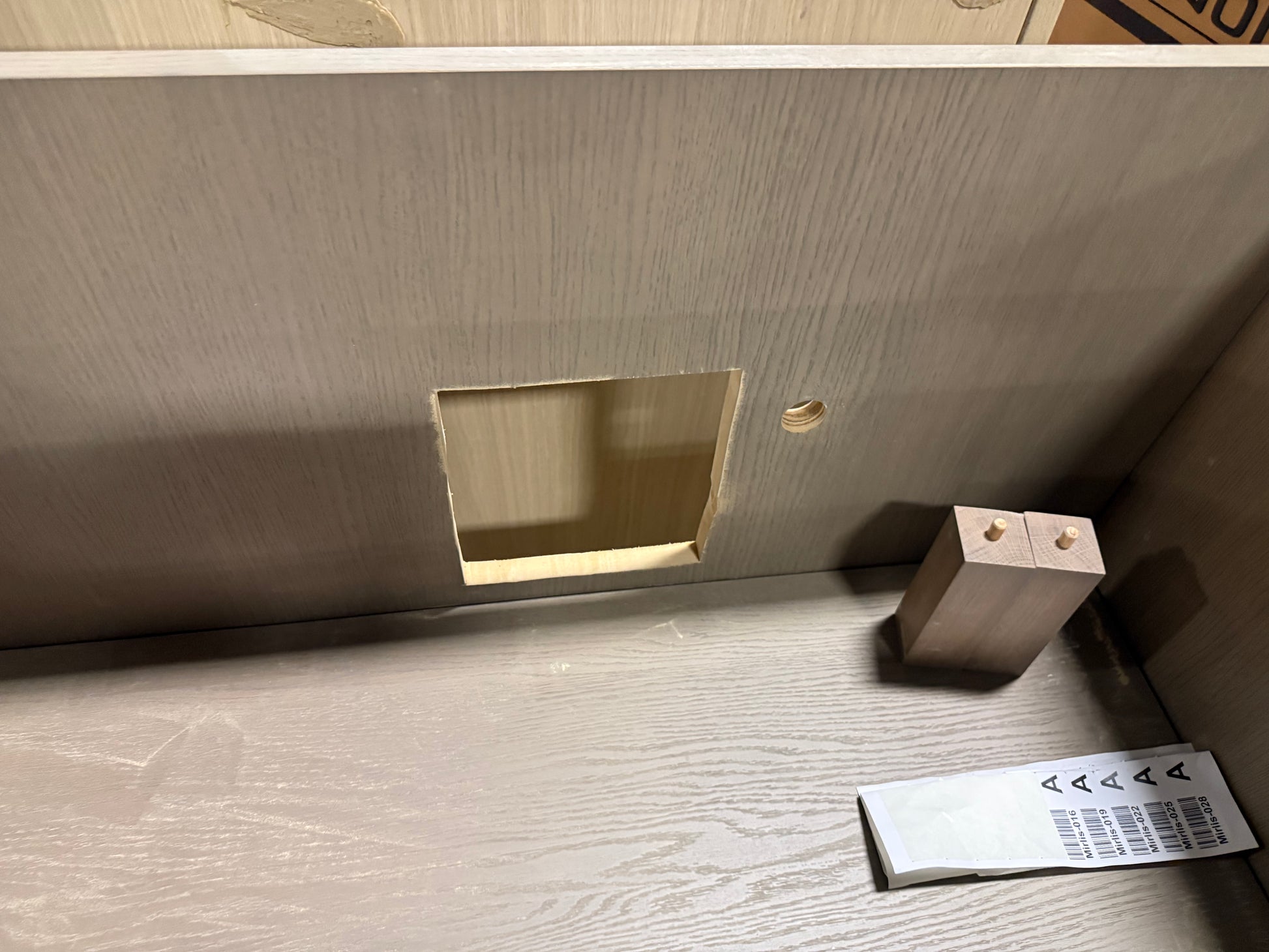 A close-up of a light wood grain furniture panel or cabinet backboard with a rectangular cutout and a small round hole, shown on a matching wood surface with a small wooden block and a paper tag nearby.