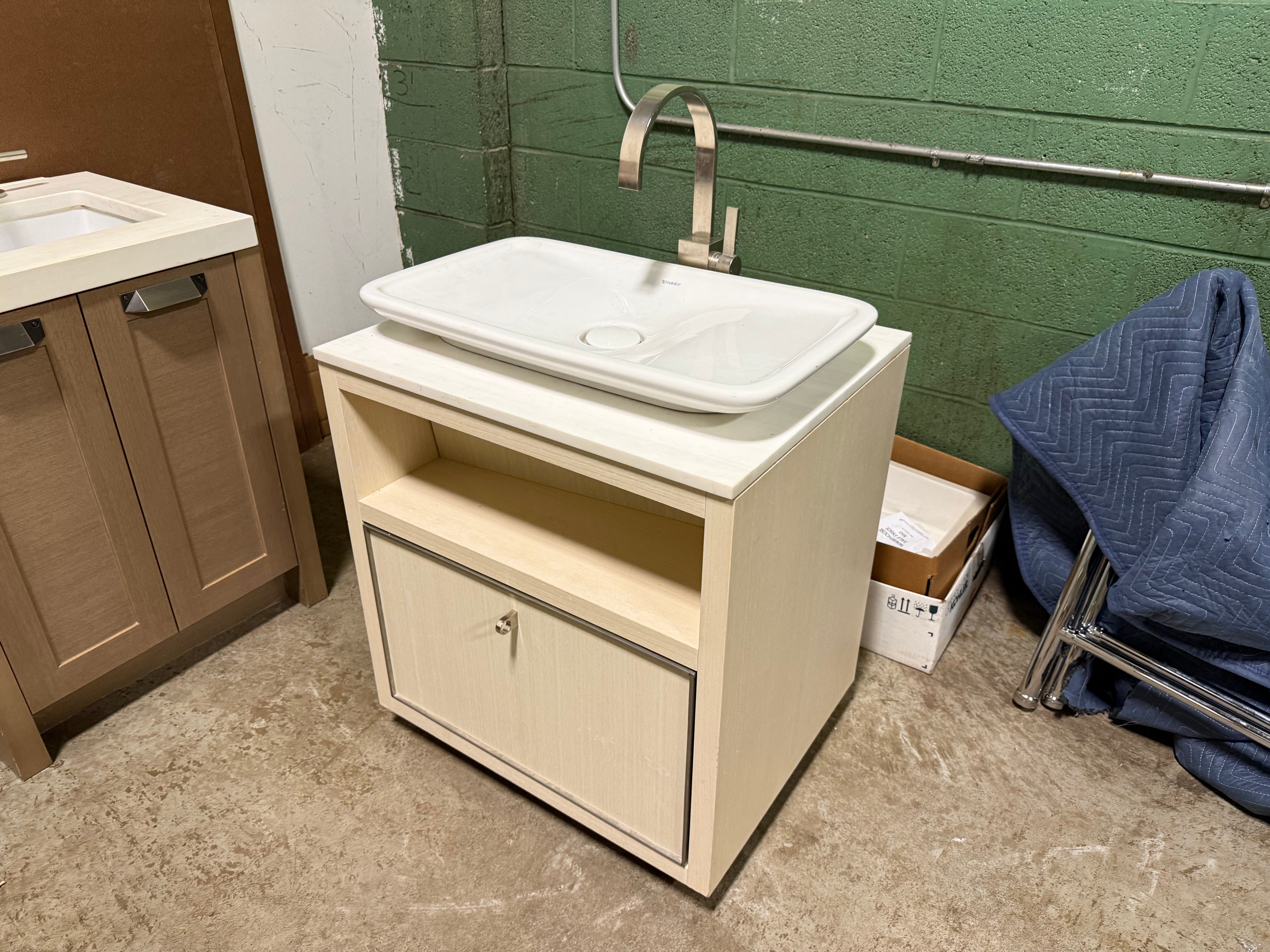 A bathroom vanity cabinet with a white rectangular vessel sink and a curved metallic faucet, shown in a warehouse or workshop setting with a green brick wall and tiled floor.