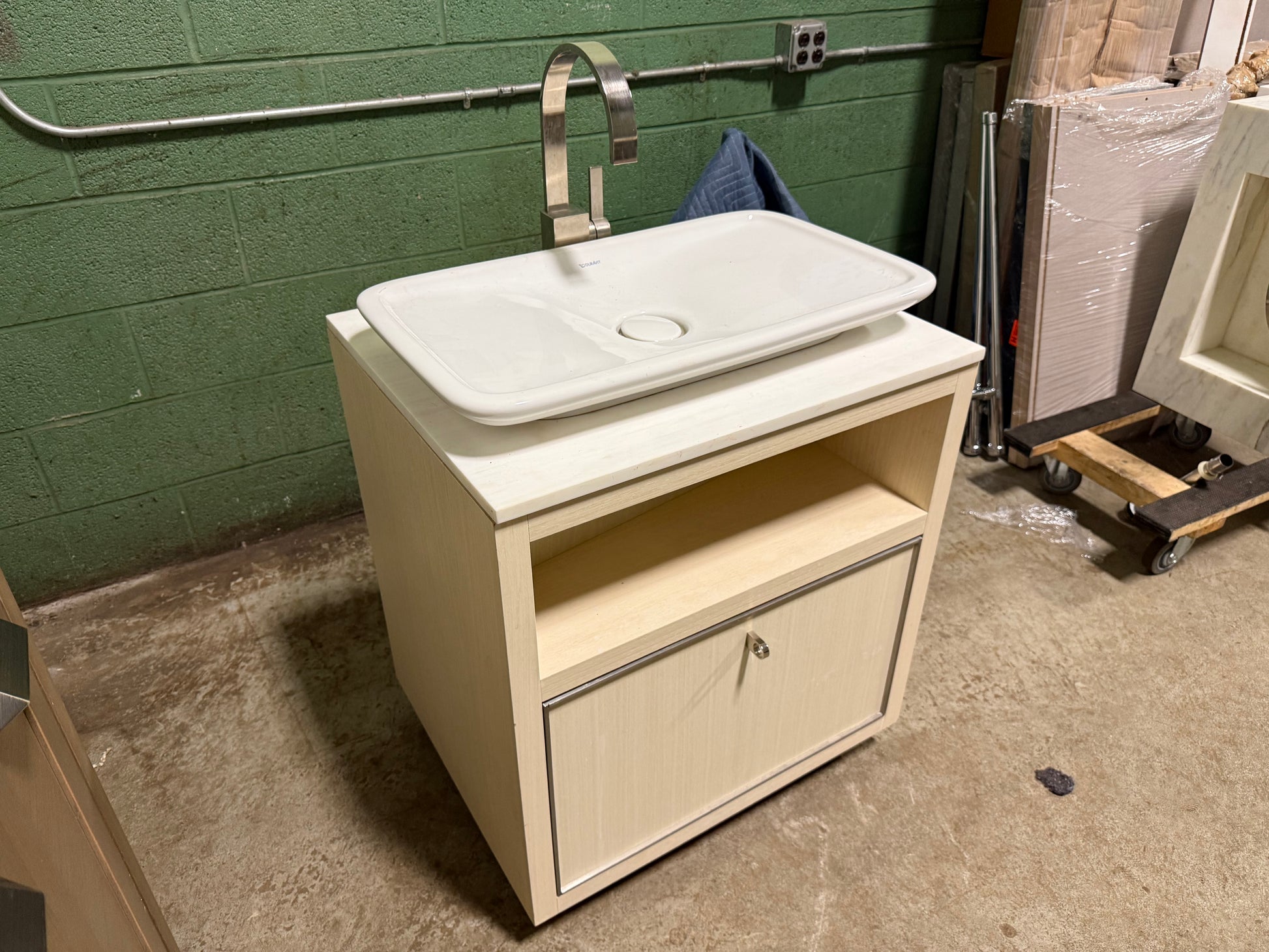 A bathroom vanity cabinet with a white rectangular vessel sink and a chrome gooseneck faucet, photographed in a warehouse or showroom setting with a green brick wall and concrete floor.