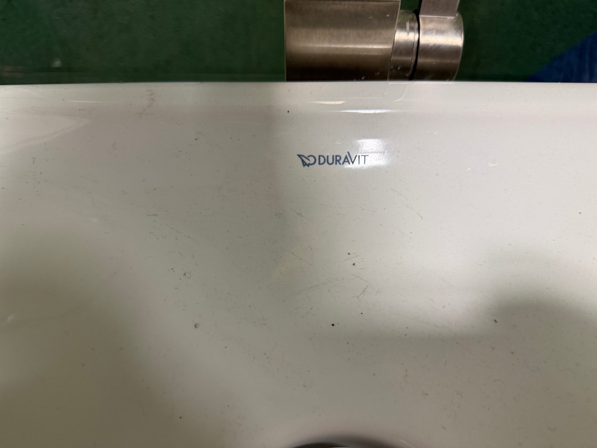 Close-up of a white Duravit sink basin with the brand logo printed in the center, showing some surface scuffs and a dark green background above.