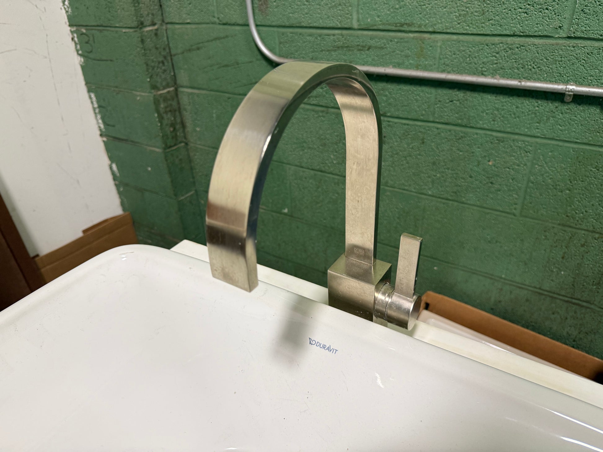 A modern curved stainless steel faucet installed on a white sink basin, set against a green painted brick wall.
