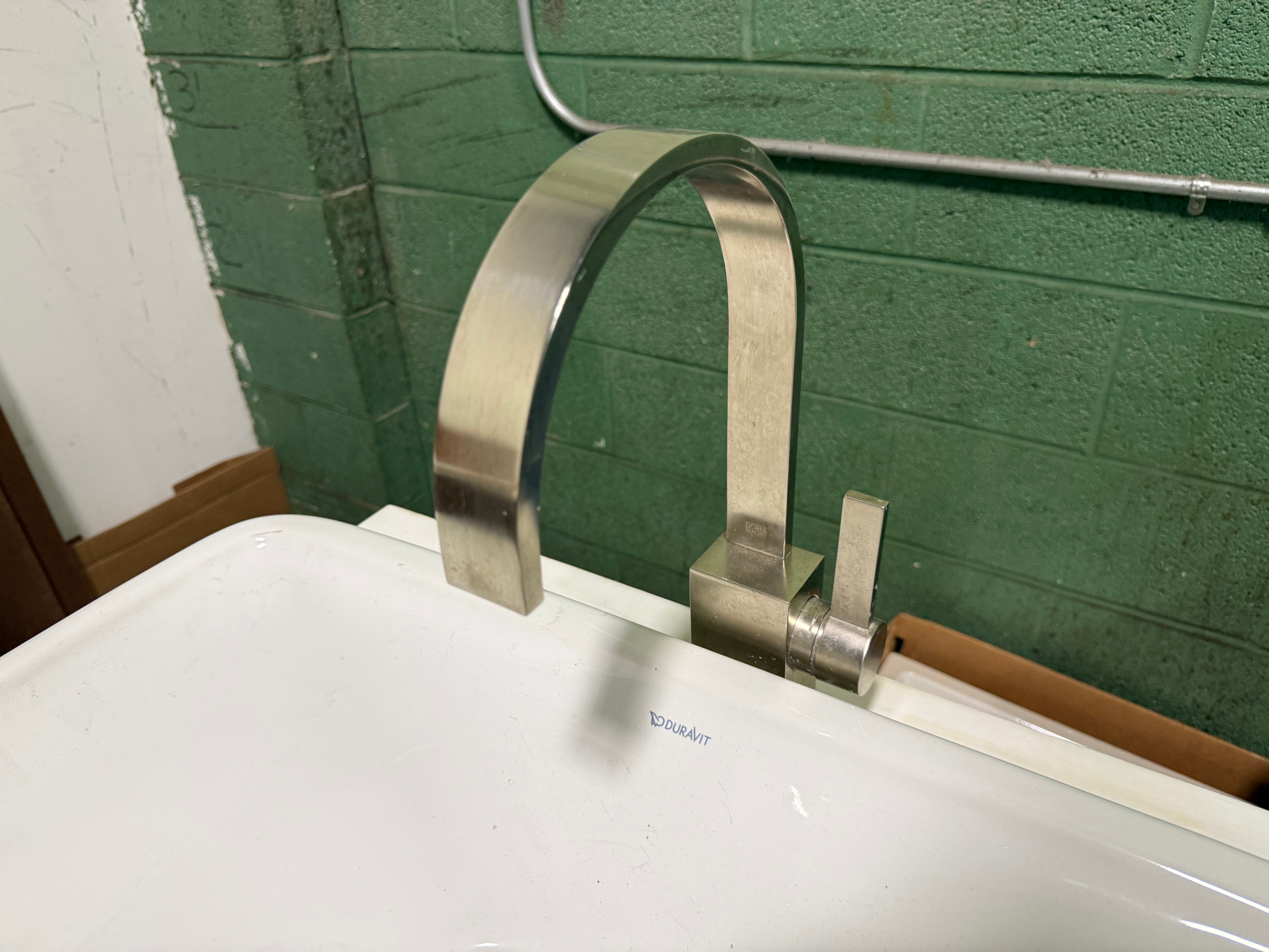 A modern curved stainless steel faucet installed on a white sink basin, set against a green painted brick wall.