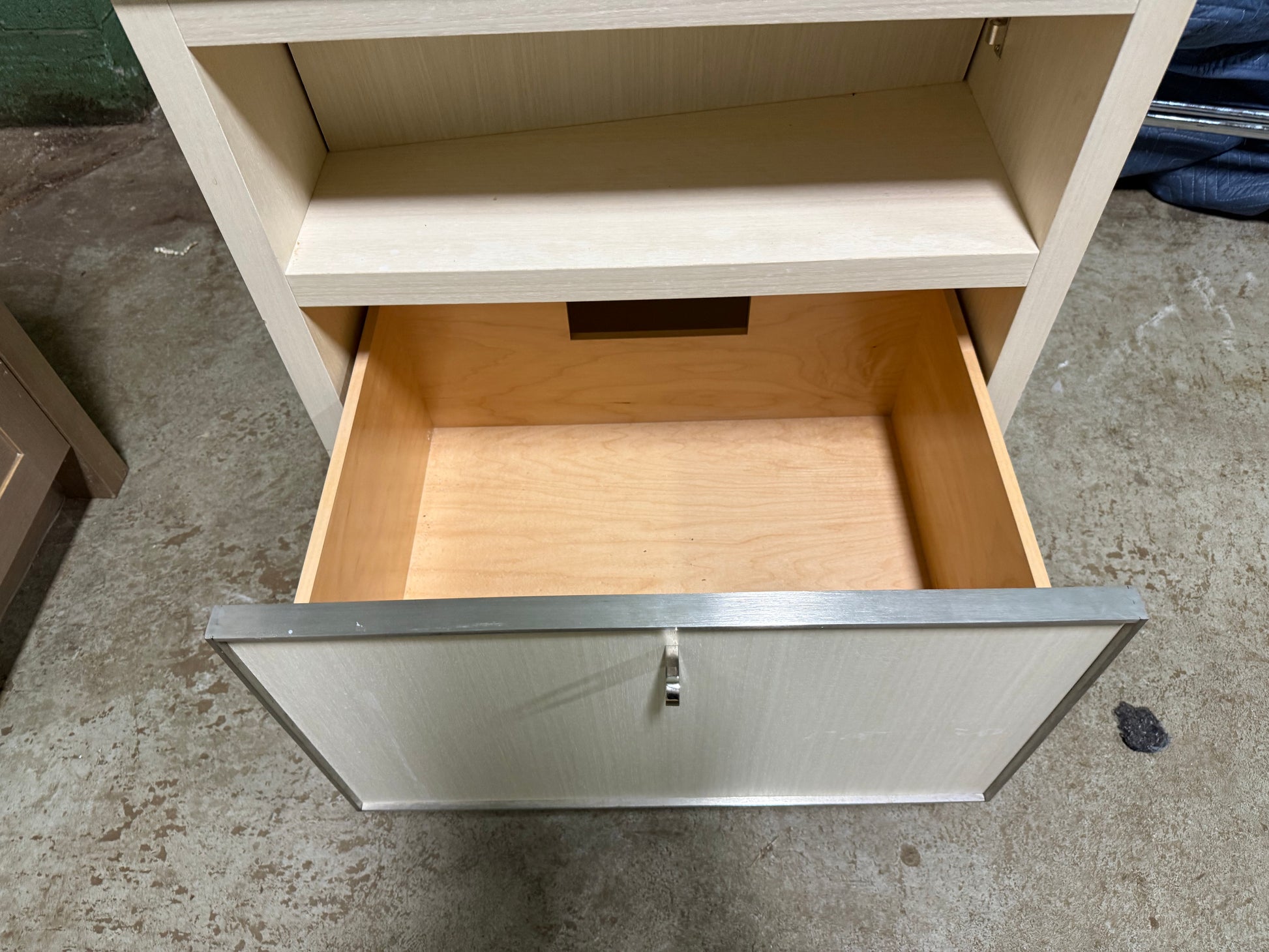 A light wood drawer cabinet with an open bottom drawer and an open upper shelf, photographed on a concrete floor.